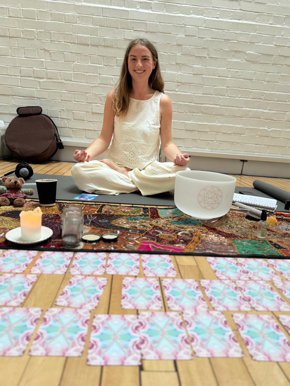 Sally, founder of Lush Lunar, sits cross-legged in a meditative pose, smiling as she holds space for ceremony in Whitley Bay. In front of her, oracle cards, a crystal singing bowl, and sacred tools create a ritual setting.